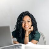 Joyful businesswoman with curly hair smiling at camera while using laptop indoors.