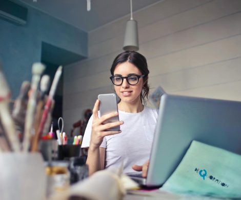 A woman using her phone at a desk, surrounded by art supplies and a laptop, in a creative workspace.